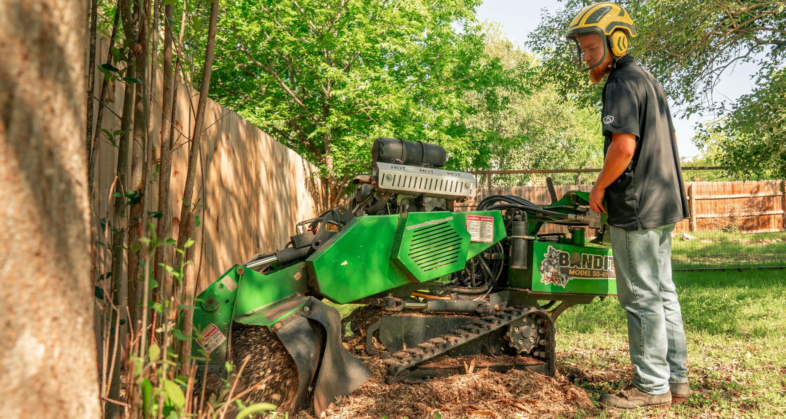 Worker operating a green stump grinder while wearing protective safety gear outdoors.